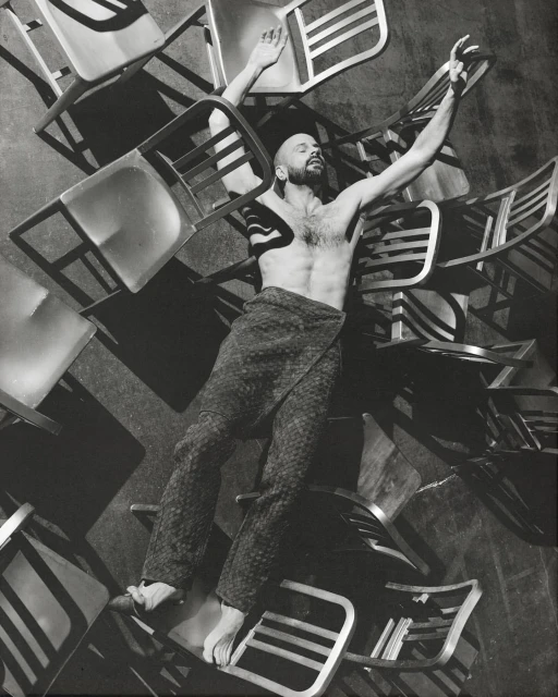 Monochrome portrait of a shirtless man lying on a pile of chairs with arms raised in an indoor setting with a dark background.