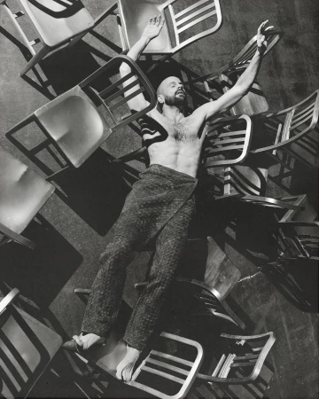 Monochrome portrait of a shirtless man lying on a pile of chairs with arms raised in an indoor setting with a dark background.