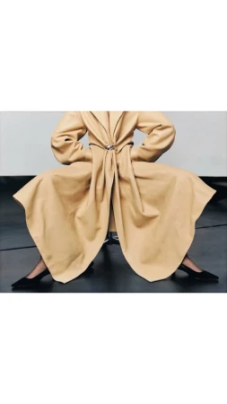 Model seated wearing a structured tan coat and black heels against a neutral studio backdrop