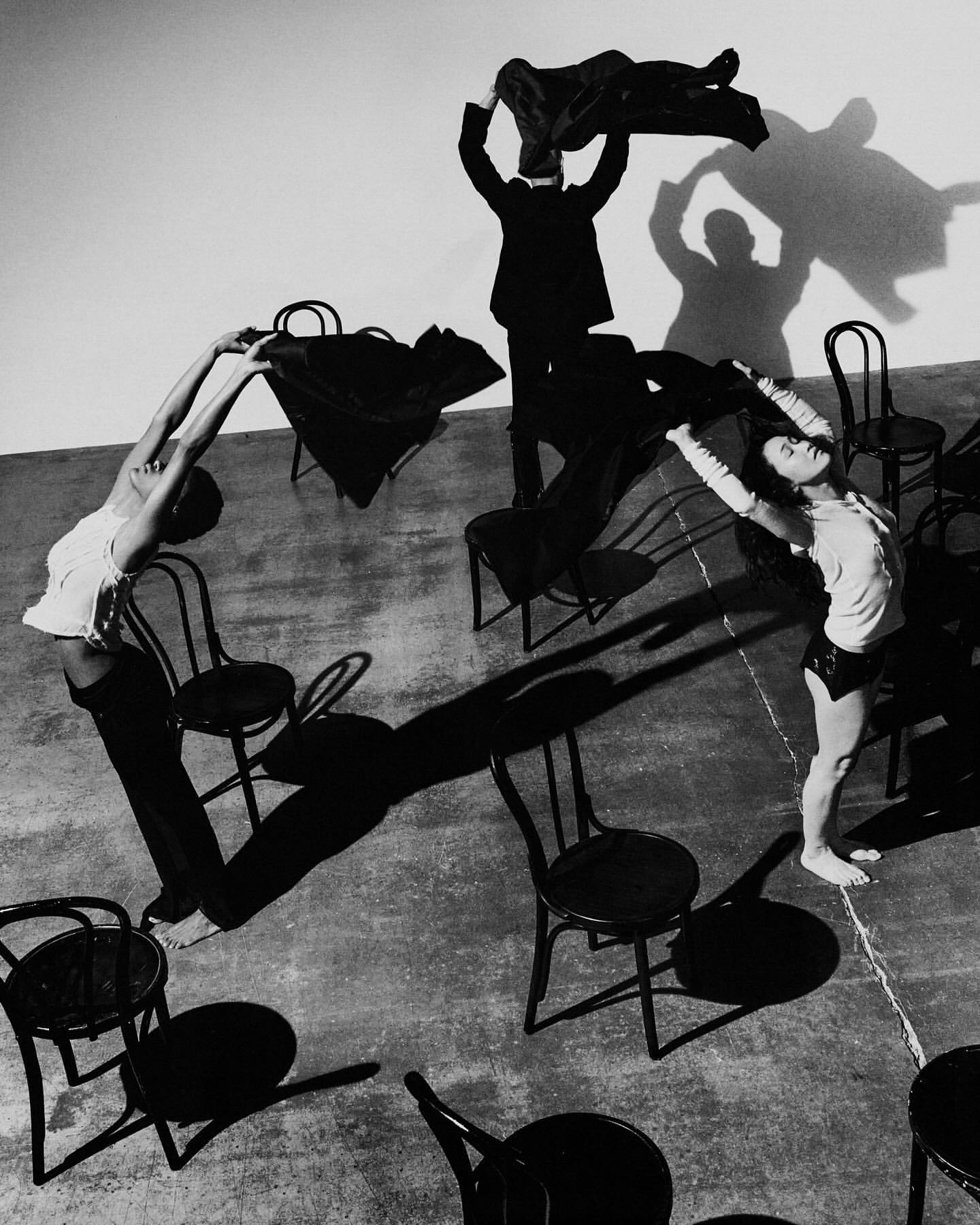 Black and white editorial photograph of three figures posed among black wooden chairs with dramatic shadows cast on a concrete floor