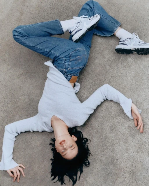 A young adult woman in blue jeans and white sneakers reclines on a gray surface, her black hair spread, and she's wearing a light sweater.