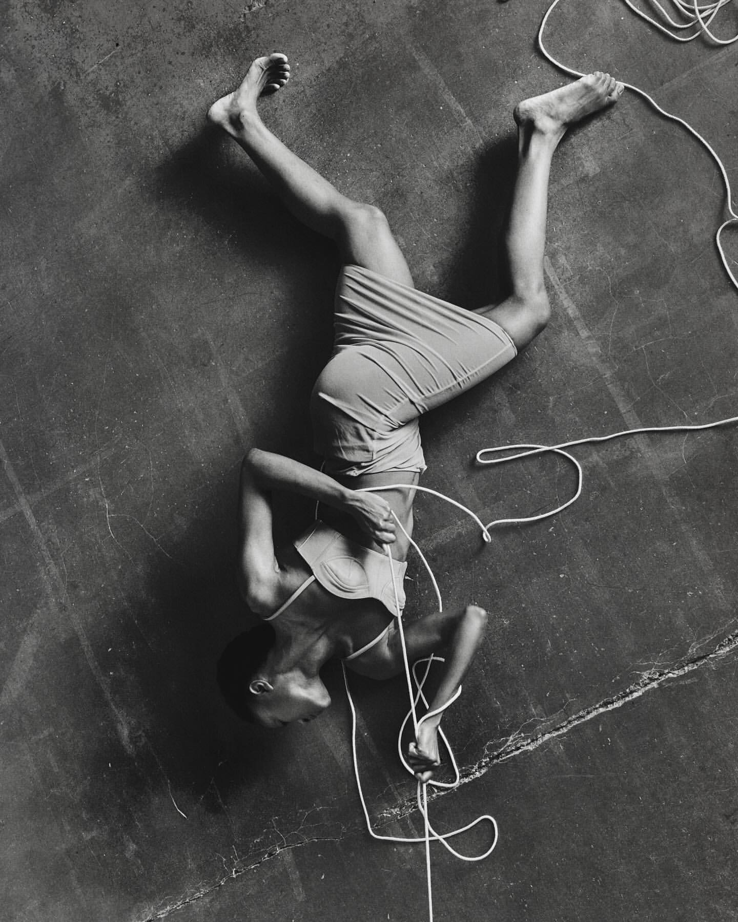 Monochrome portrait of a person lying on their back on a gray surface tangled in white rope, shot at a high angle.