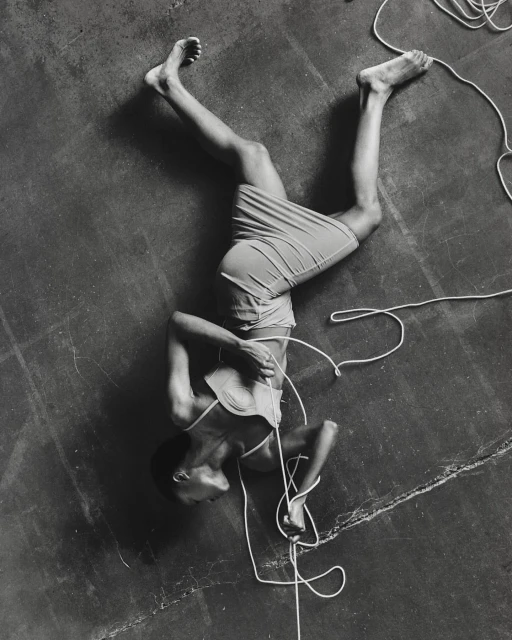 Monochrome portrait of a person lying on their back on a gray surface tangled in white rope, shot at a high angle.
