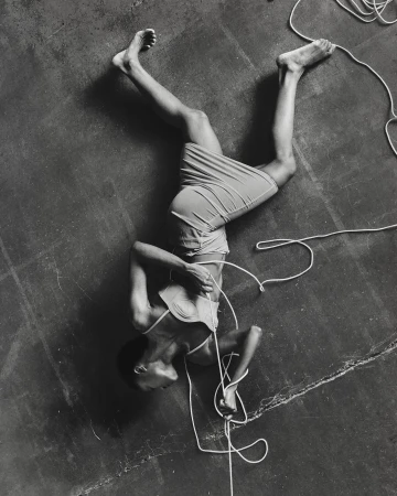 Monochrome portrait of a person lying on their back on a gray surface tangled in white rope, shot at a high angle.