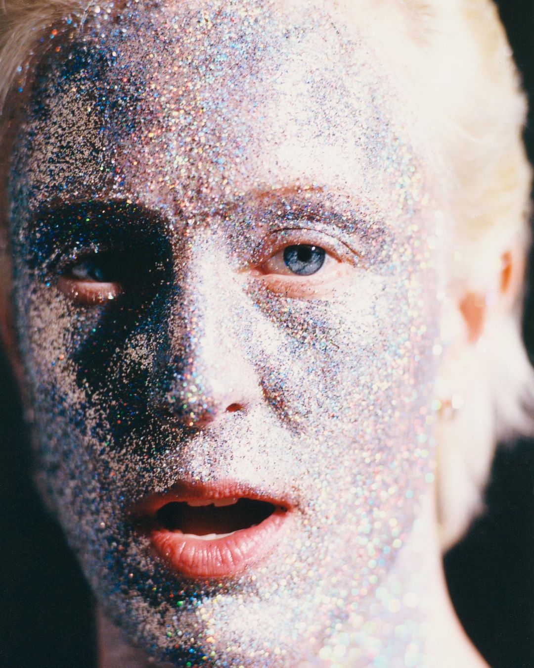 Close-up portrait of an adult woman covered in silver glitter on her face, fair skin, blue eyes, light blonde hair, against a dark backdrop.