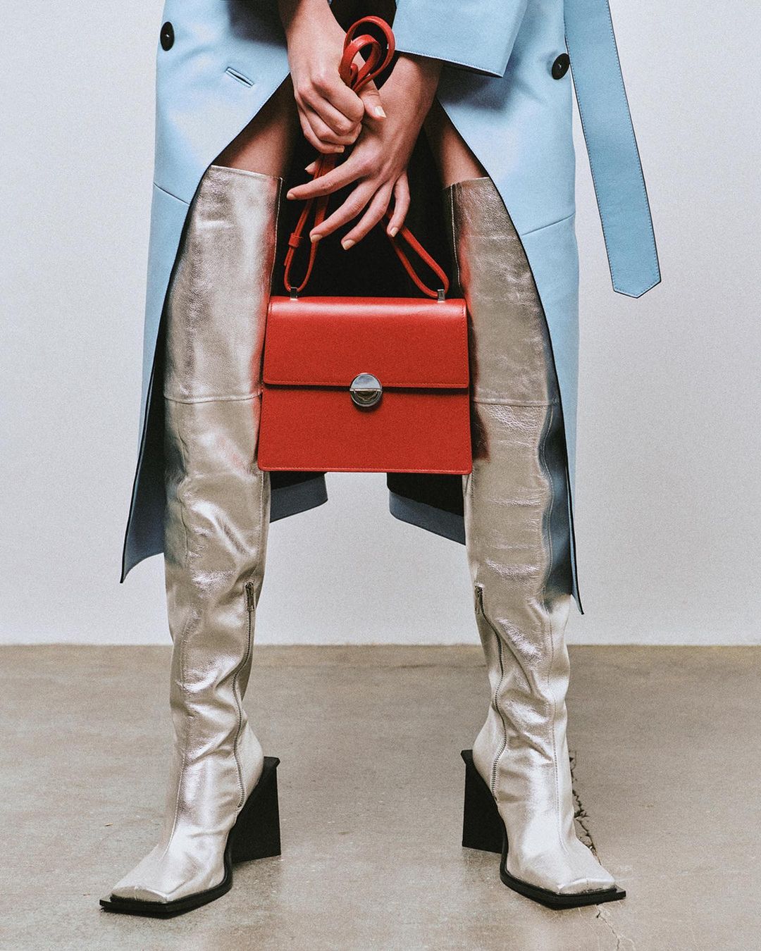Fashion shot featuring silver thigh-high boots, red handbag, light blue trench coat, held by fair-skinned hands against a neutral backdrop.