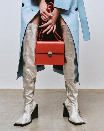 Fashion shot featuring silver thigh-high boots, red handbag, light blue trench coat, held by fair-skinned hands against a neutral backdrop.