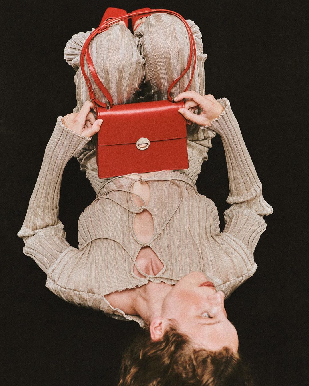 Fashion portrait of a young adult female lying down in a gray ribbed top holding a small red leather purse with a black background.