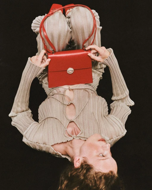 Fashion portrait of a young adult female lying down in a gray ribbed top holding a small red leather purse with a black background.