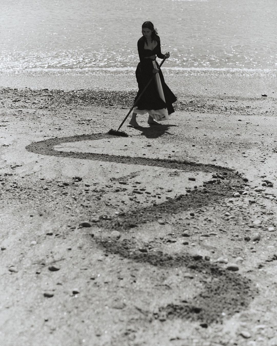 Monochrome shot of a woman raking sand at the beach, creating a swirling design in the sand near the ocean waters.