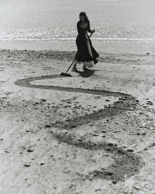 Monochrome shot of a woman raking sand at the beach, creating a swirling design in the sand near the ocean waters.