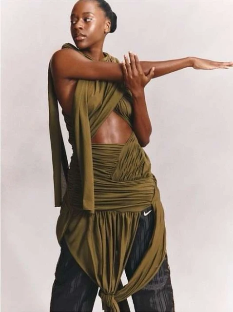 Adult woman poses in a draped olive-green dress with bare midriff and black athletic pants in a neutral indoor setting.