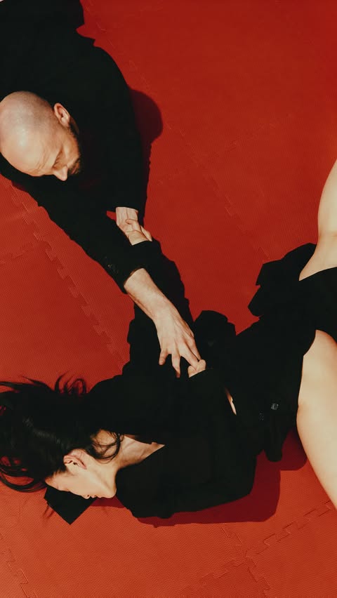 A woman and a bald man are lying on a red wrestling mat, both wearing black, with the man pinning the woman from an overhead perspective.
