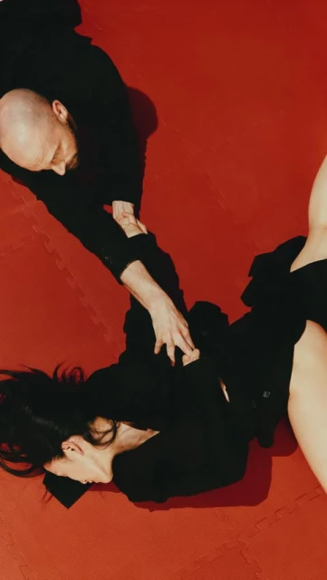 A woman and a bald man are lying on a red wrestling mat, both wearing black, with the man pinning the woman from an overhead perspective.