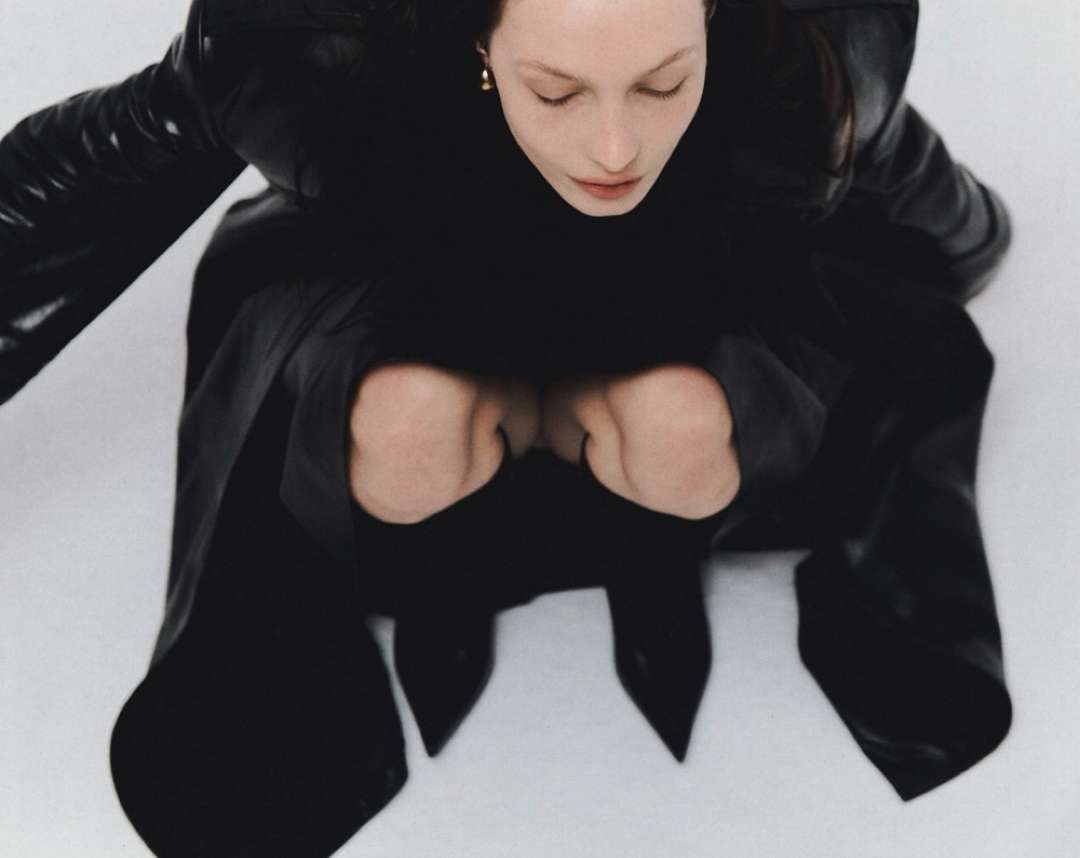 Woman in black outfit squats on white background in a high-angle shot, creating a minimalist, fashion-forward image.