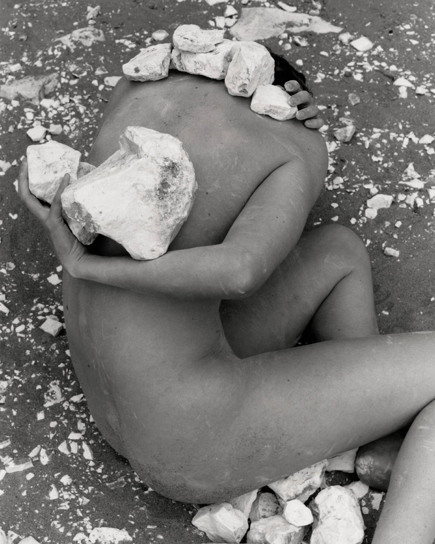 Monochrome photograph of an adult lying on sand, cradling large rocks, with smaller stones balanced on head in an outdoor setting.