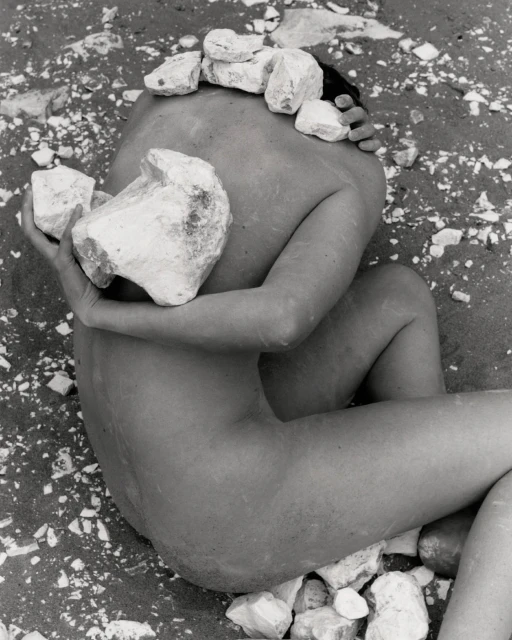 Monochrome photograph of an adult lying on sand, cradling large rocks, with smaller stones balanced on head in an outdoor setting.