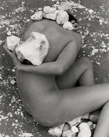 Monochrome photograph of an adult lying on sand, cradling large rocks, with smaller stones balanced on head in an outdoor setting.