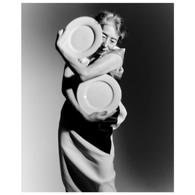 Monochrome portrait of a woman hugging two white plates, with a dark and light background and wearing a black and white dress.