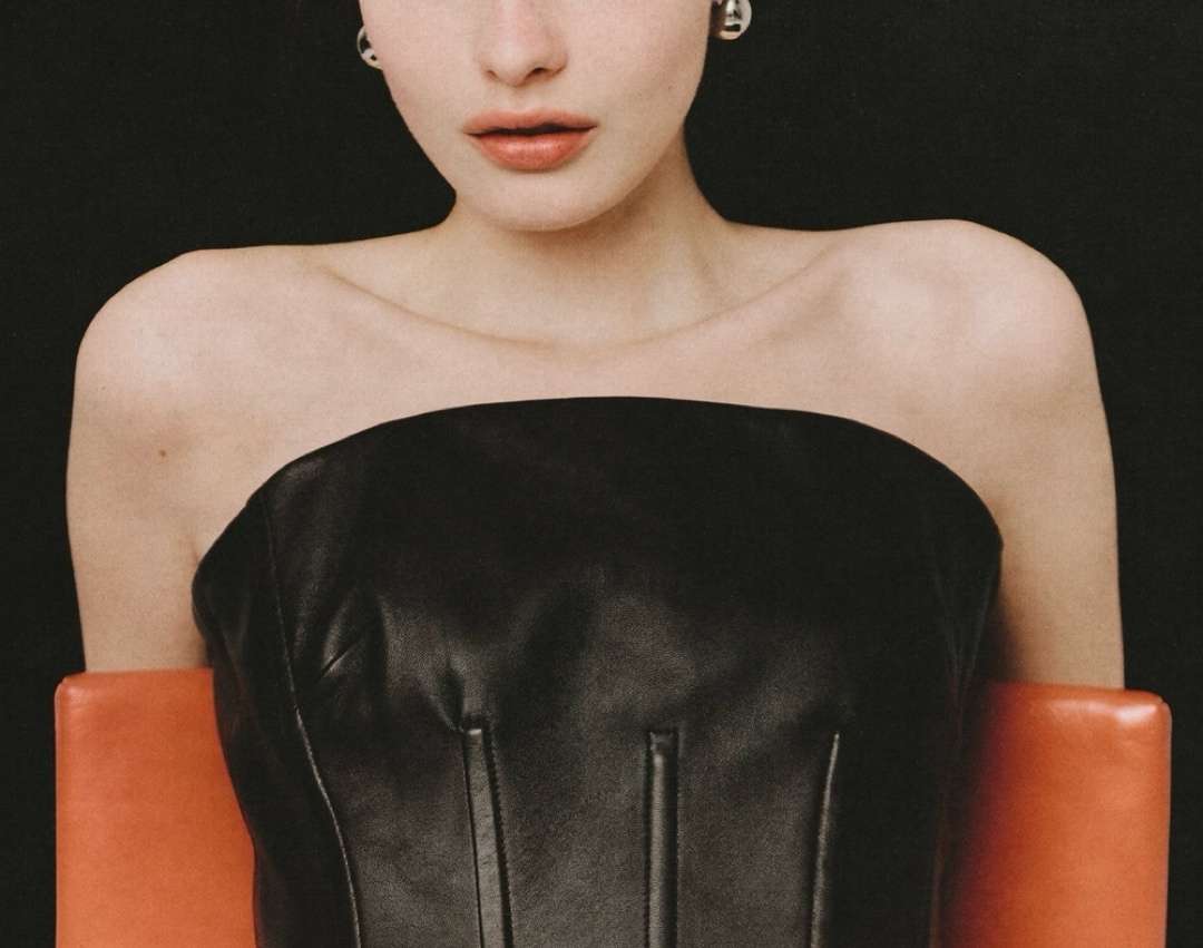 Close-up studio shot of a fair-skinned young adult woman wearing a strapless black top, seated on a orange chair against a black background.