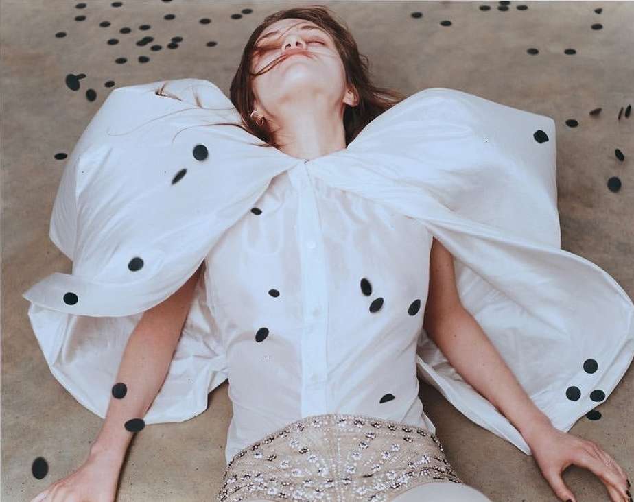 Fashion shot of a model lying on the ground in a white shirt with large sleeves, embellished shorts, and black polka dots scattered around.