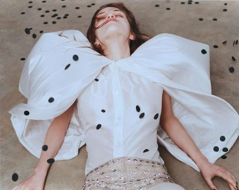 Fashion shot of a model lying on the ground in a white shirt with large sleeves, embellished shorts, and black polka dots scattered around.