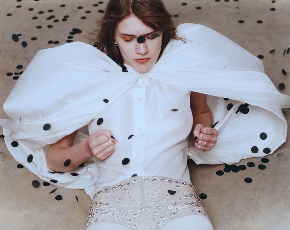 Fashion editorial shot of a woman in a white dress with black polka dots lying on a speckled surface. Dark makeup accentuates the eyes.
