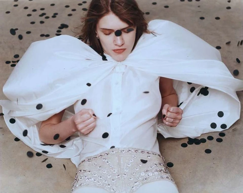 Fashion editorial shot of a woman in a white dress with black polka dots lying on a speckled surface. Dark makeup accentuates the eyes.