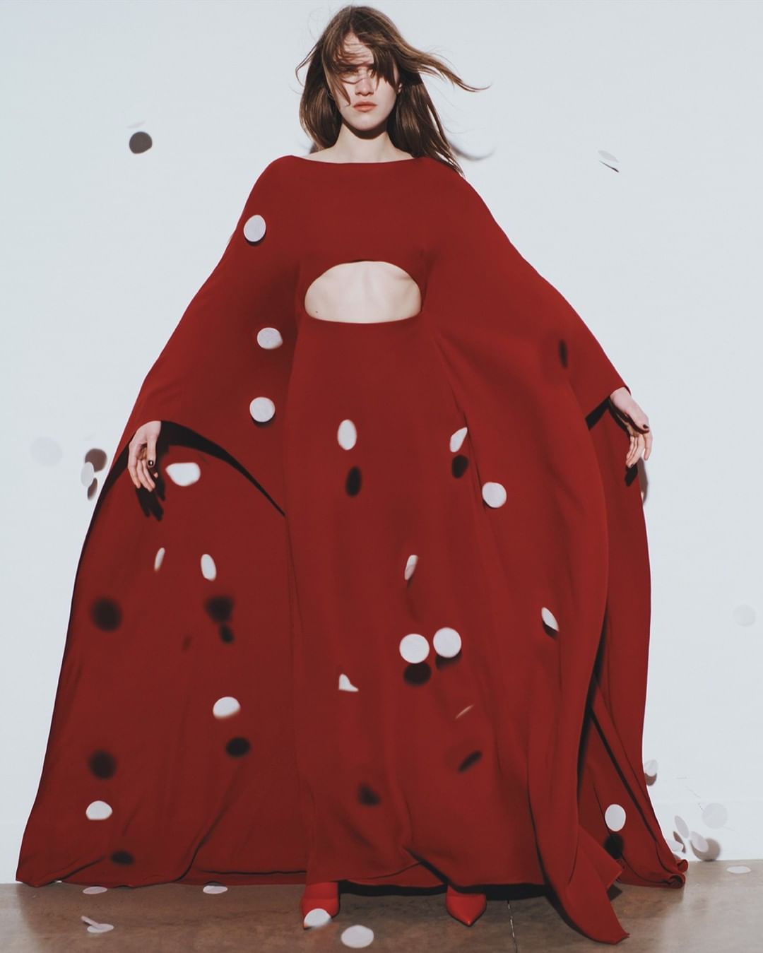 A person in a long red gown with white polka dots poses in a studio surrounded by falling confetti. Fashion and style visual.
