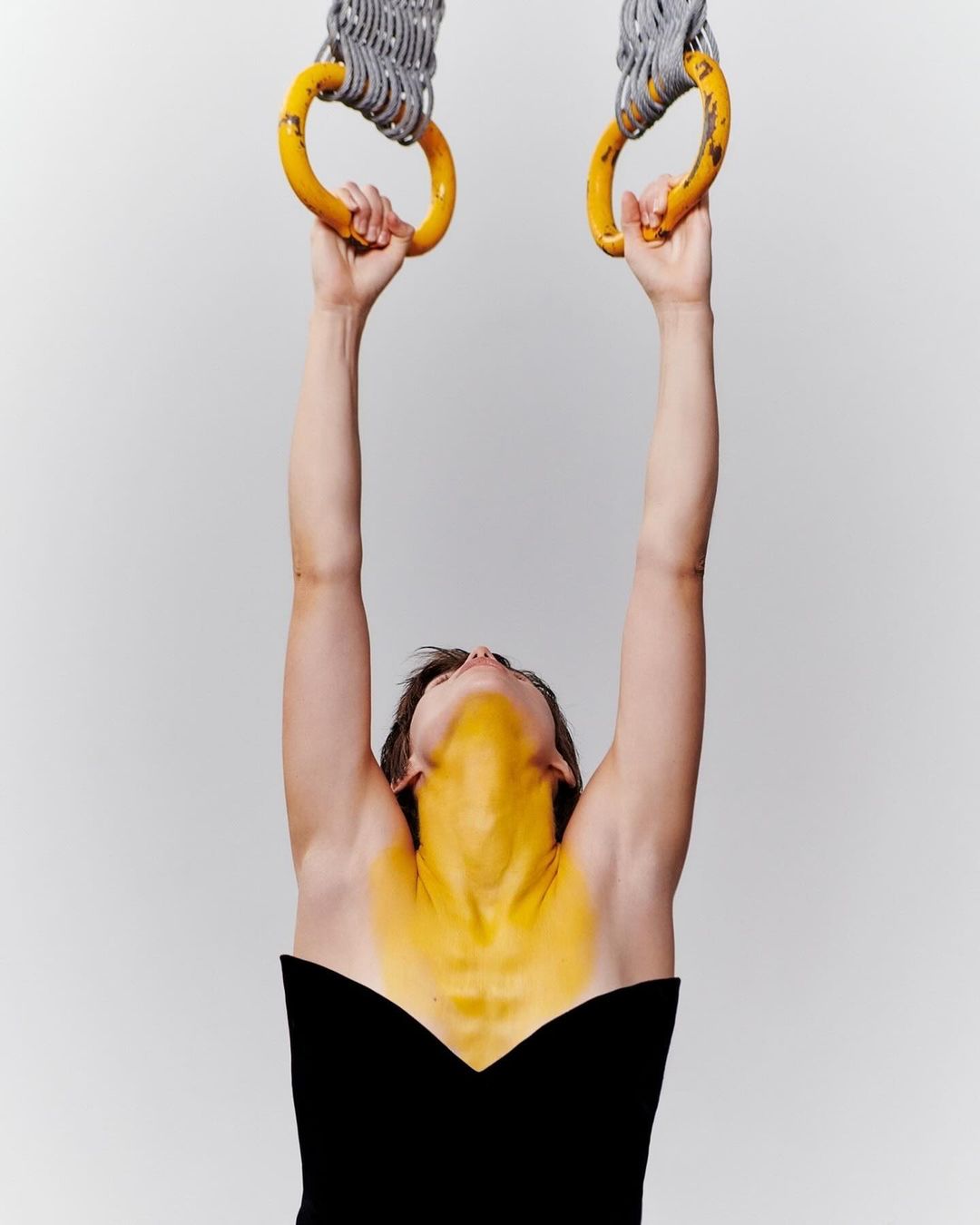 An athletic adult woman hangs from yellow gymnastics rings, shot from a low angle in a photo studio against a light-gray backdrop.