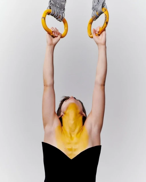 An athletic adult woman hangs from yellow gymnastics rings, shot from a low angle in a photo studio against a light-gray backdrop.
