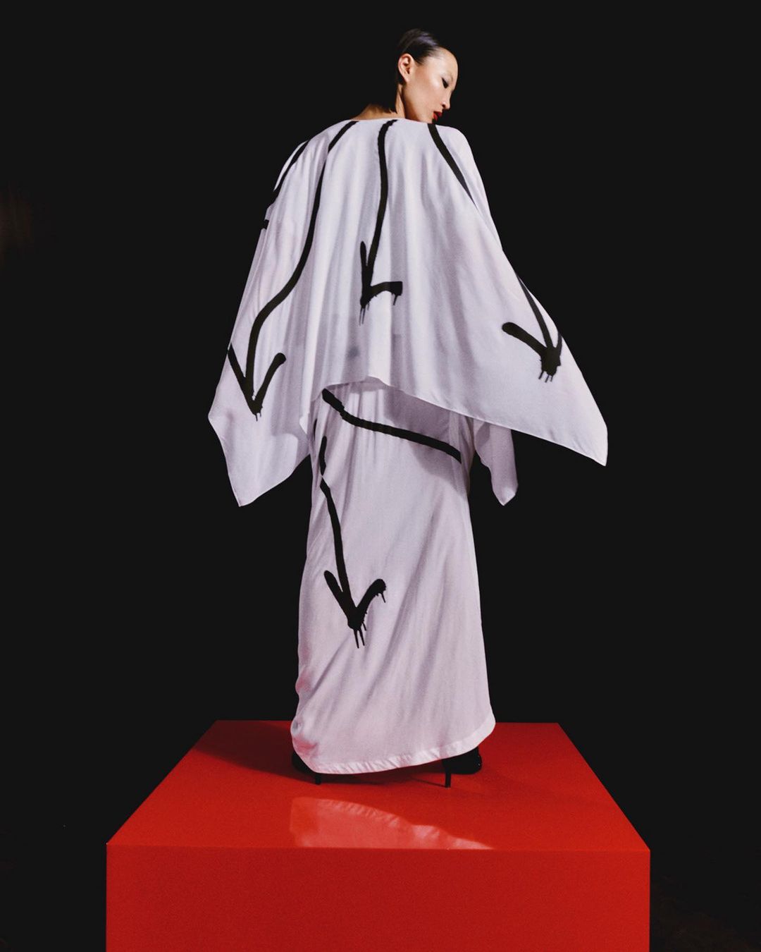 A fashion shot presents a woman wearing a white outfit with black arrow designs, standing on a red platform against a black background.