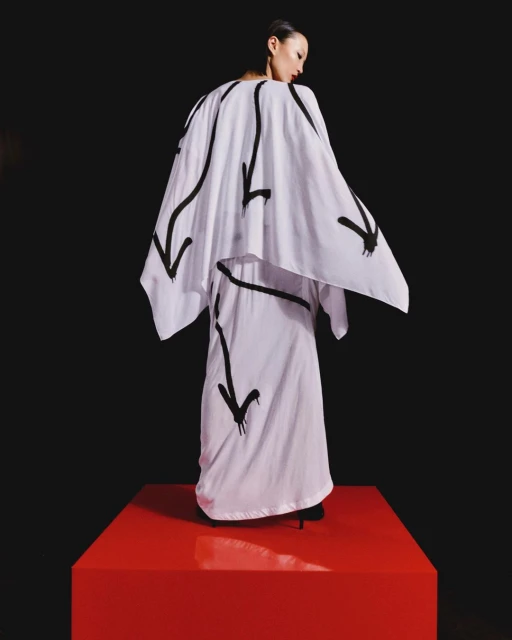 A fashion shot presents a woman wearing a white outfit with black arrow designs, standing on a red platform against a black background.