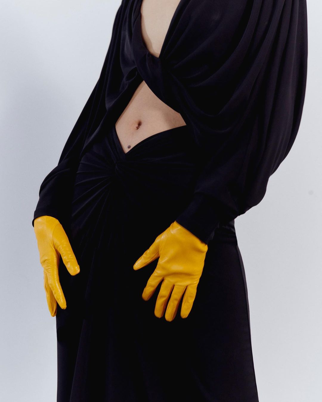 Woman in a black dress with a midriff cut-out, posing with yellow leather gloves against a white background, fashion studio shot.