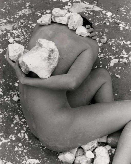 Black and white image of a nude human form lying on a sandy surface covered with white stones, some resting directly on the body.