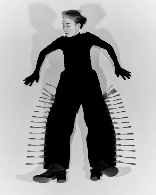 An adult female lies on a white surface wearing black clothing and shoes, surrounded by a series of forks framing the body.