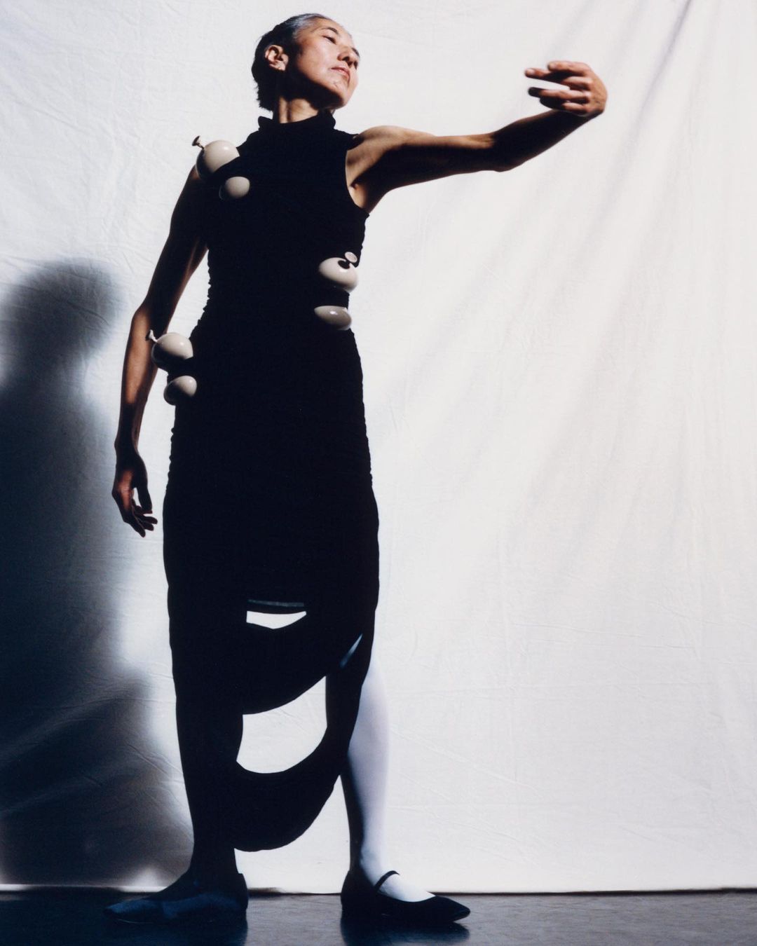 Full shot of an Asian woman with short gray hair, wearing a black dress with white spheres attached, striking a modern dance pose in studio.