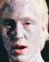Close-up portrait of a person with silver glitter covering their face and skin against a dark background