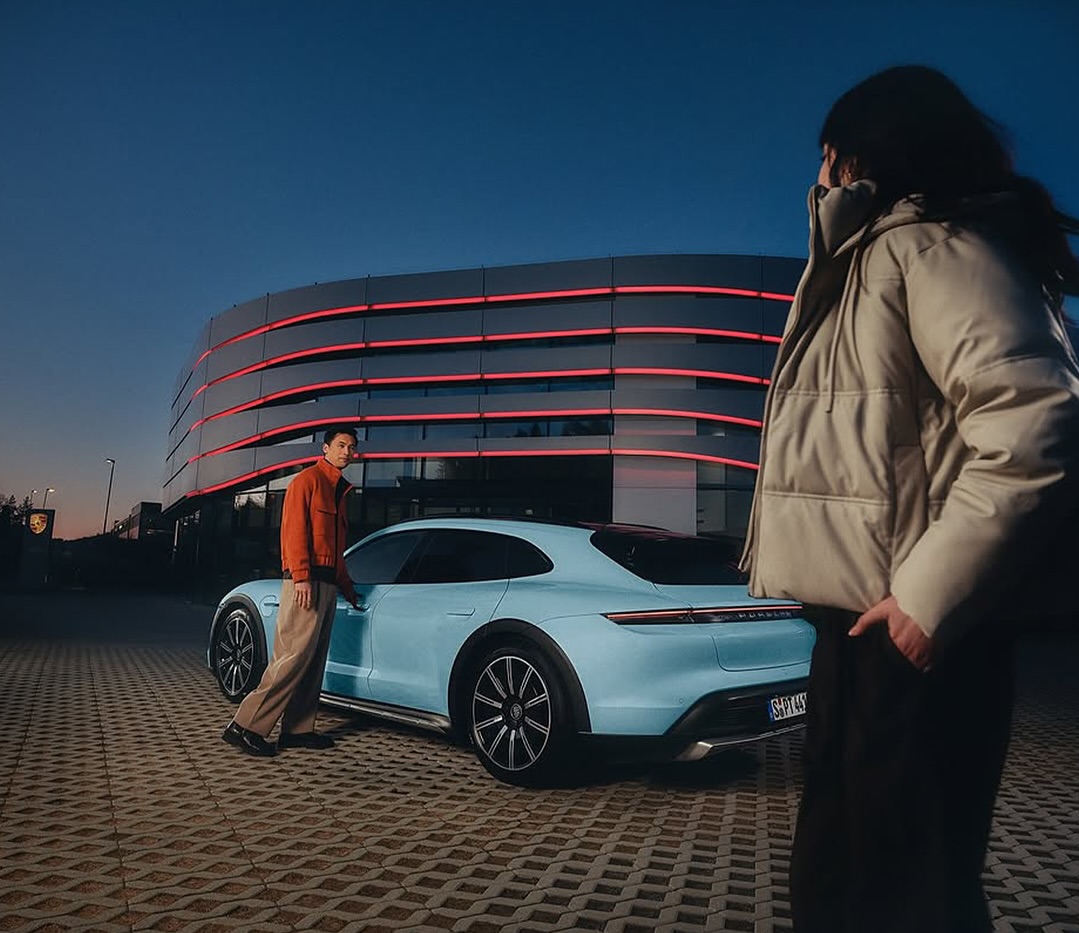 Two people stand near a light-blue Porsche Taycan Cross Turismo in front of an illuminated modern building against a dusk sky.