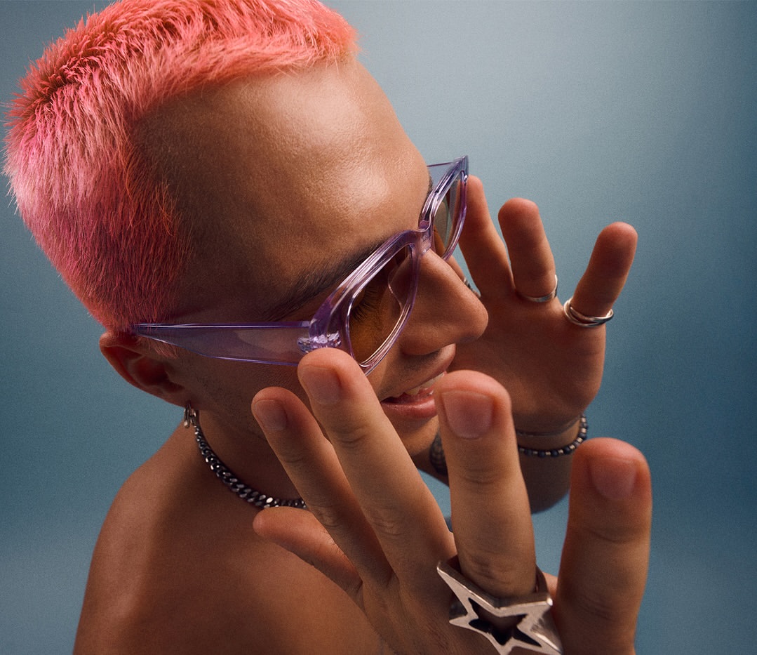 Portrait of a person with short pink hair wearing translucent purple sunglasses and silver rings by Lara Baumberger