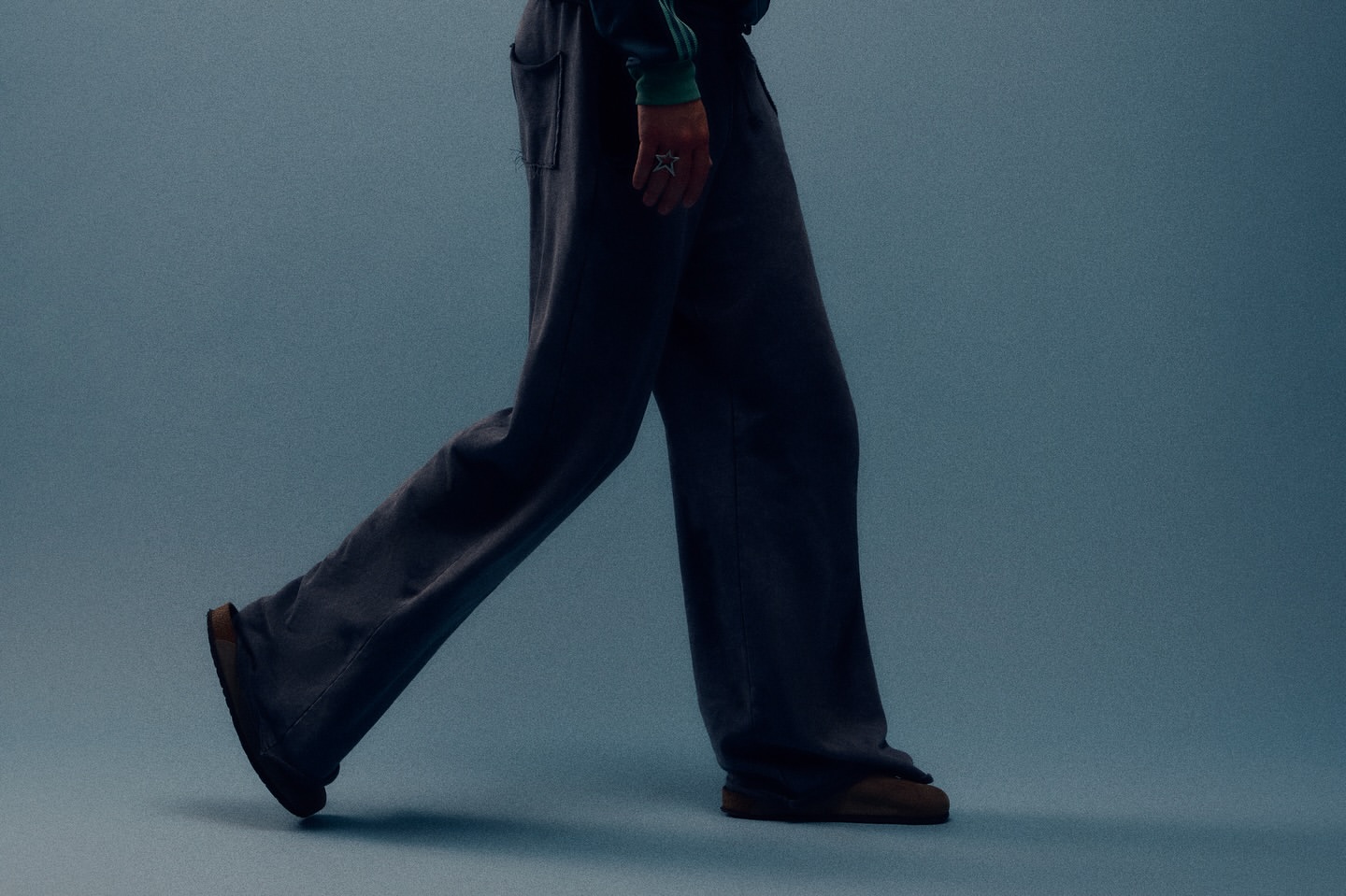 A person walking with gray sweatpants and brown shoes against a blue background with soft lighting, creating a relaxed and casual visual.