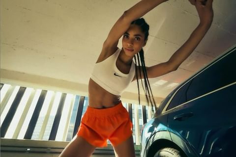 Fit woman stretching in orange shorts and sports bra next to a car in a parking garage with sunlight streaming through window slats.