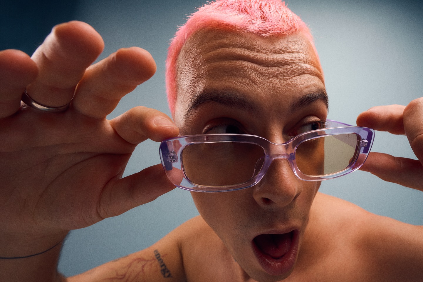 Close-up shot of a young adult with pink hair wearing lilac glasses, making a surprised facial expression against a blue backdrop.