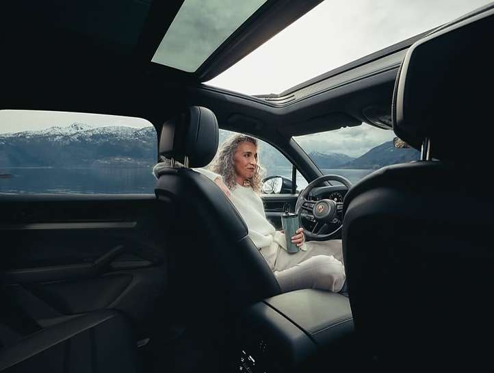 Woman sits in a Porsche SUV by a lake, snow-capped mountains visible through the sunroof and side windows. Car interior in focus.