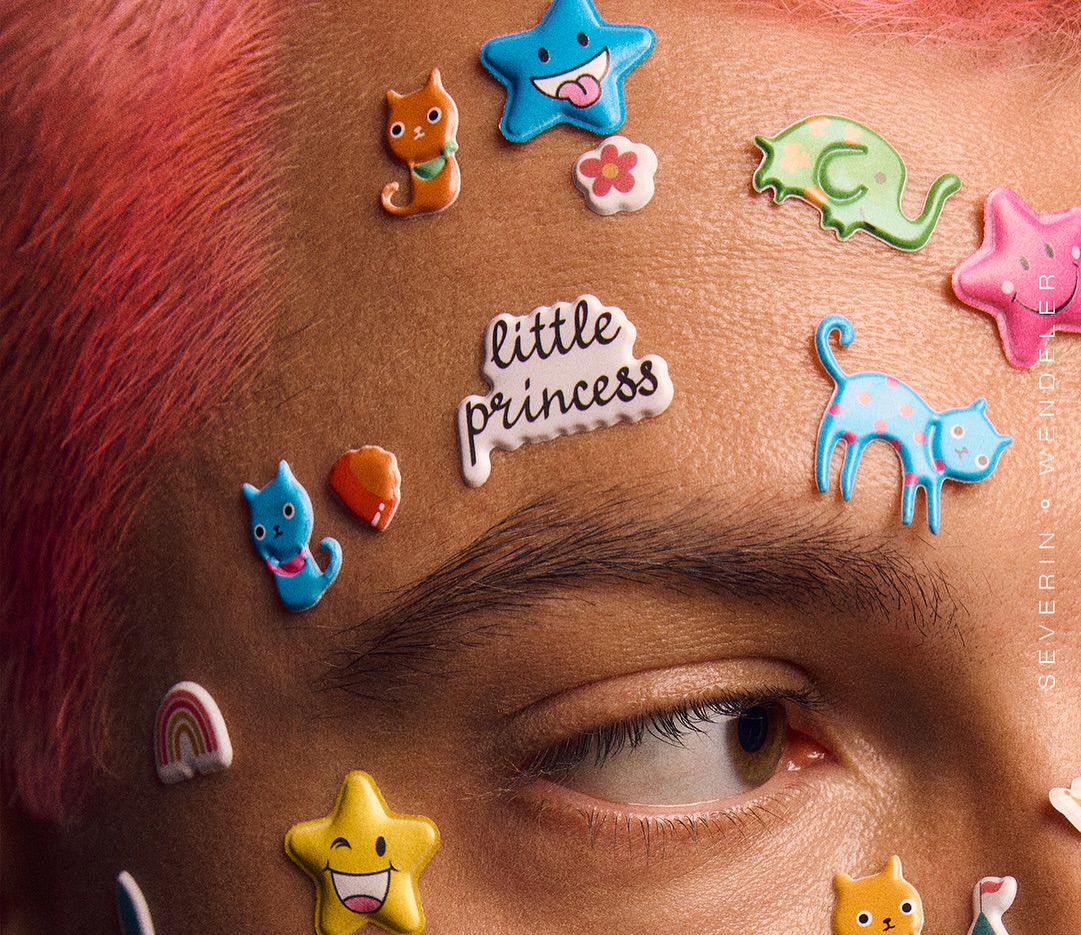 Close-up of a person with pink hair and stickers on their forehead, including stars, cats, a rainbow, and the text 'little princess'.