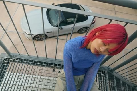 Eye-level shot of a person with vibrant red hair resting on metal stairs, with a light-colored car visible in the background.