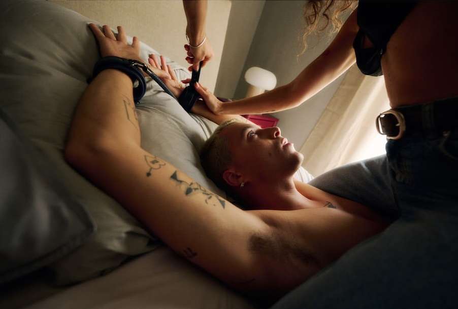 A fair-skinned adult male lies on a gray pillow while a person fastens black wrist restraints on him in an indoor bedroom setting.
