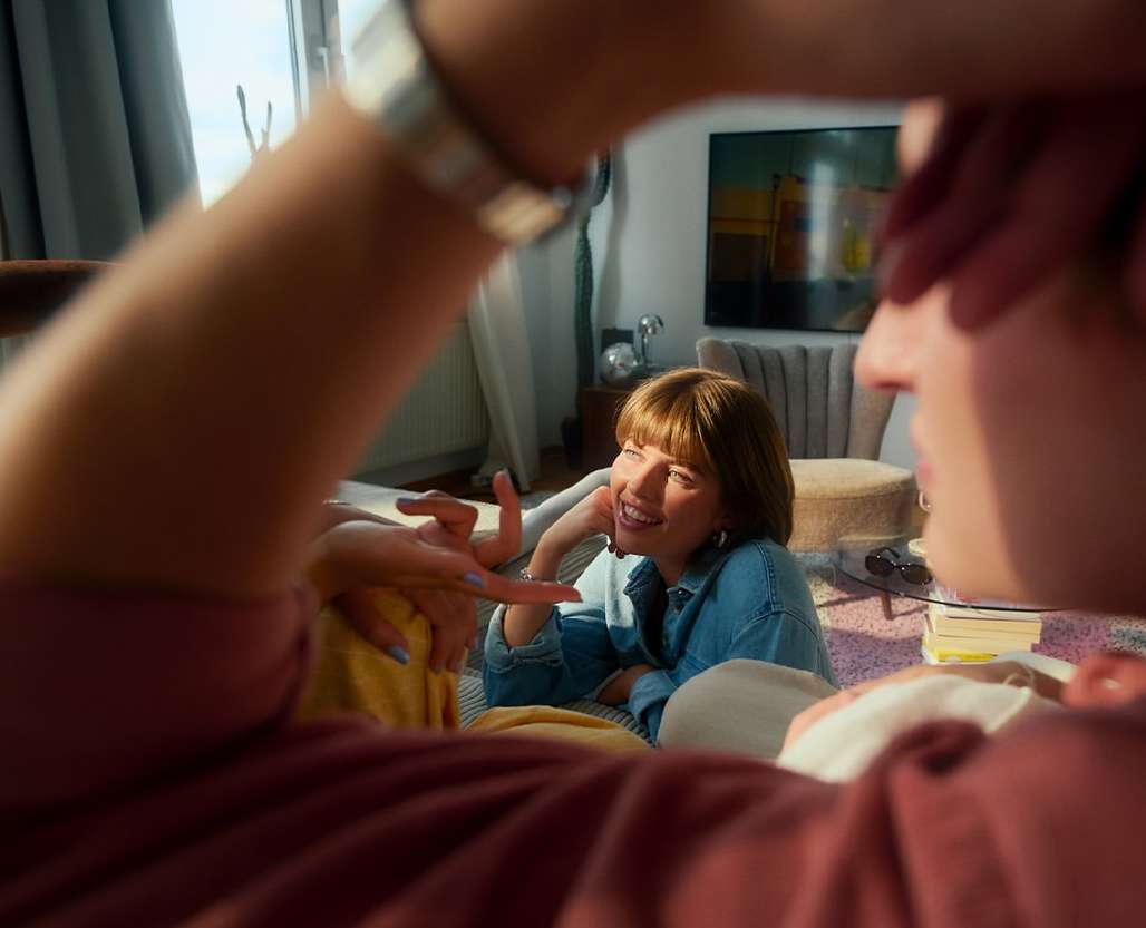 Two young adult women are indoors sitting on the floor and smiling while talking with another person, with sunlight coming through windows.