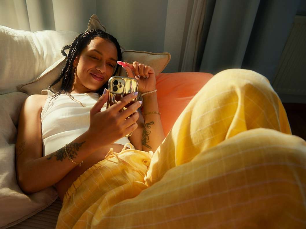 Young woman smiles while using a smartphone indoors. She is lying down wearing a tank top and yellow pants. Natural light.