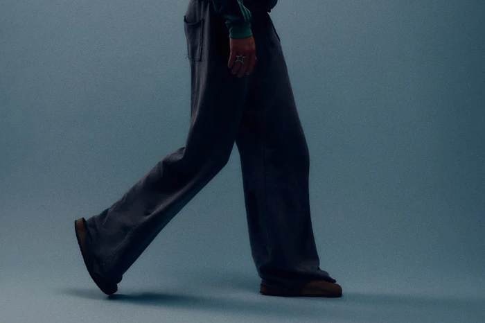 Low-angle side profile of a person walking in loose dark trousers and brown slippers against a blue studio backdrop.